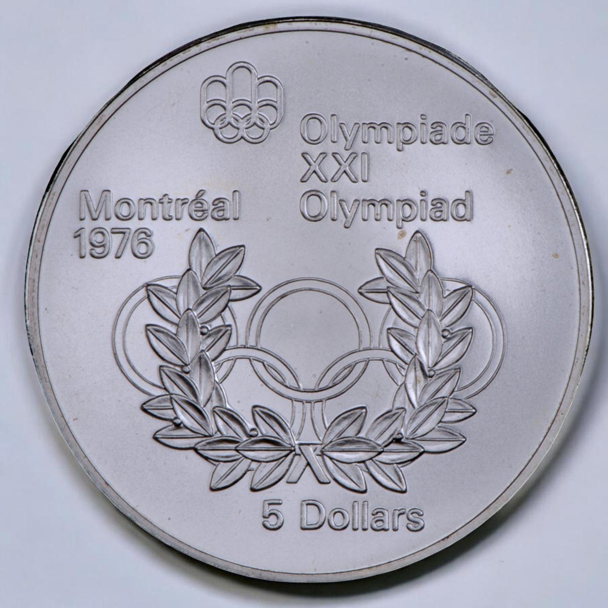Coin, silver, Olympic symbol of 3 linked rings above 2 linked rings, a wreath of leaves in front.