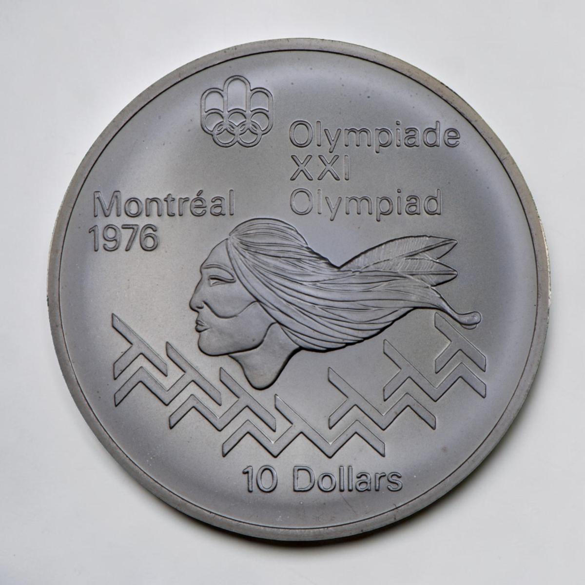 Coin, silver, Indigenous person’s head in profile above a motif of interconnected “T” shapes.
