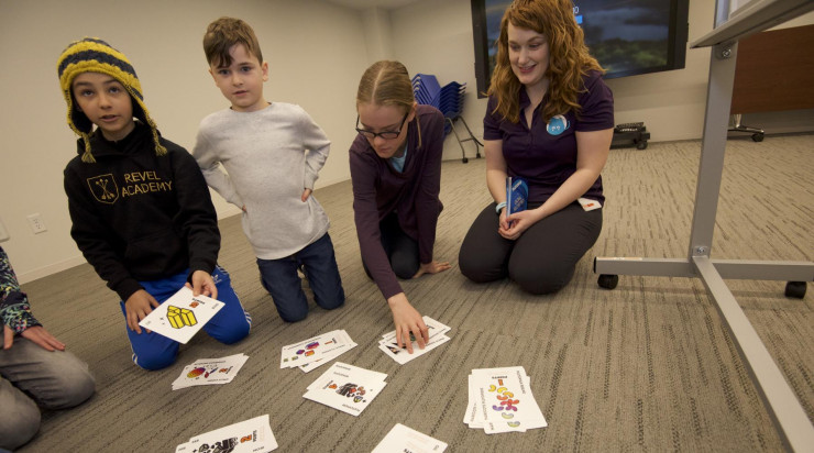 Young students sitting with their teacher on the floor looking at resource cards they will trade as part of the activity.