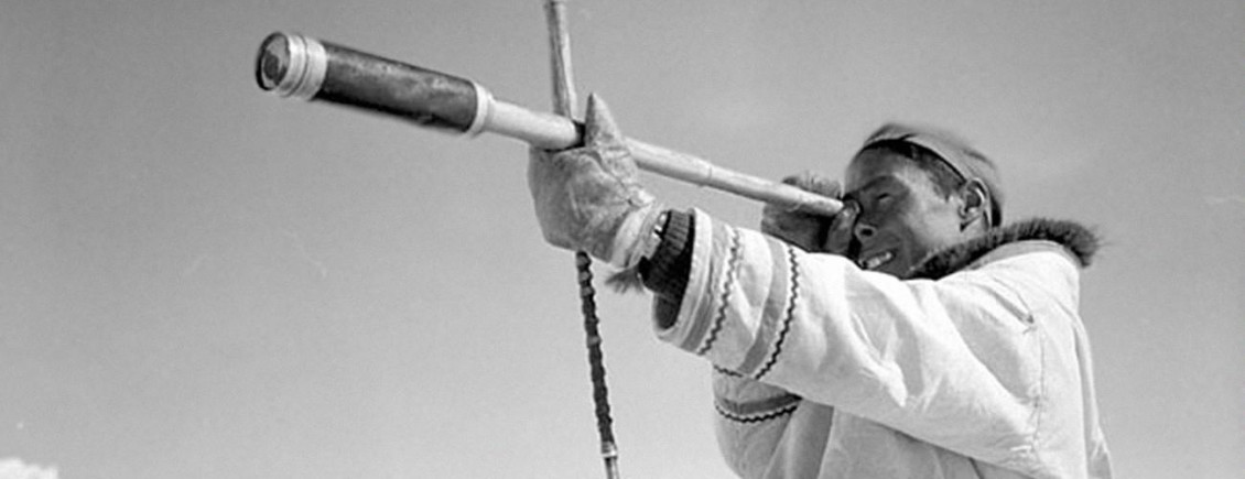 Black and white photo of an Inuit hunter looking through telescope.