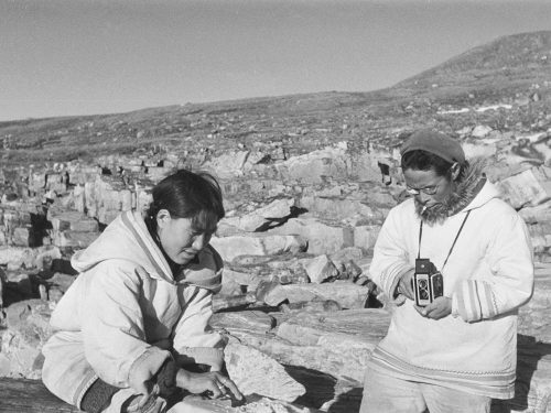 Black and white photo of Inuit man photographing a woman scraping a hide.