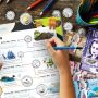 Hands filling out a form on a table with art supplies and paper play money.