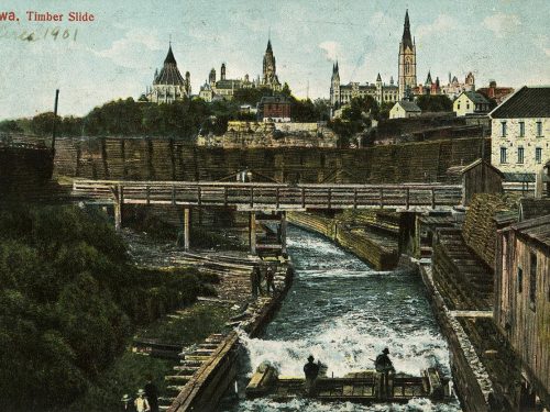 Photograph, men on a raft of squared logs ride down a canal with gothic buildings in the background.