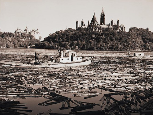 Photo, black and white, thousands of logs and two boats on a river in front of a hill with gothic towers and trees.