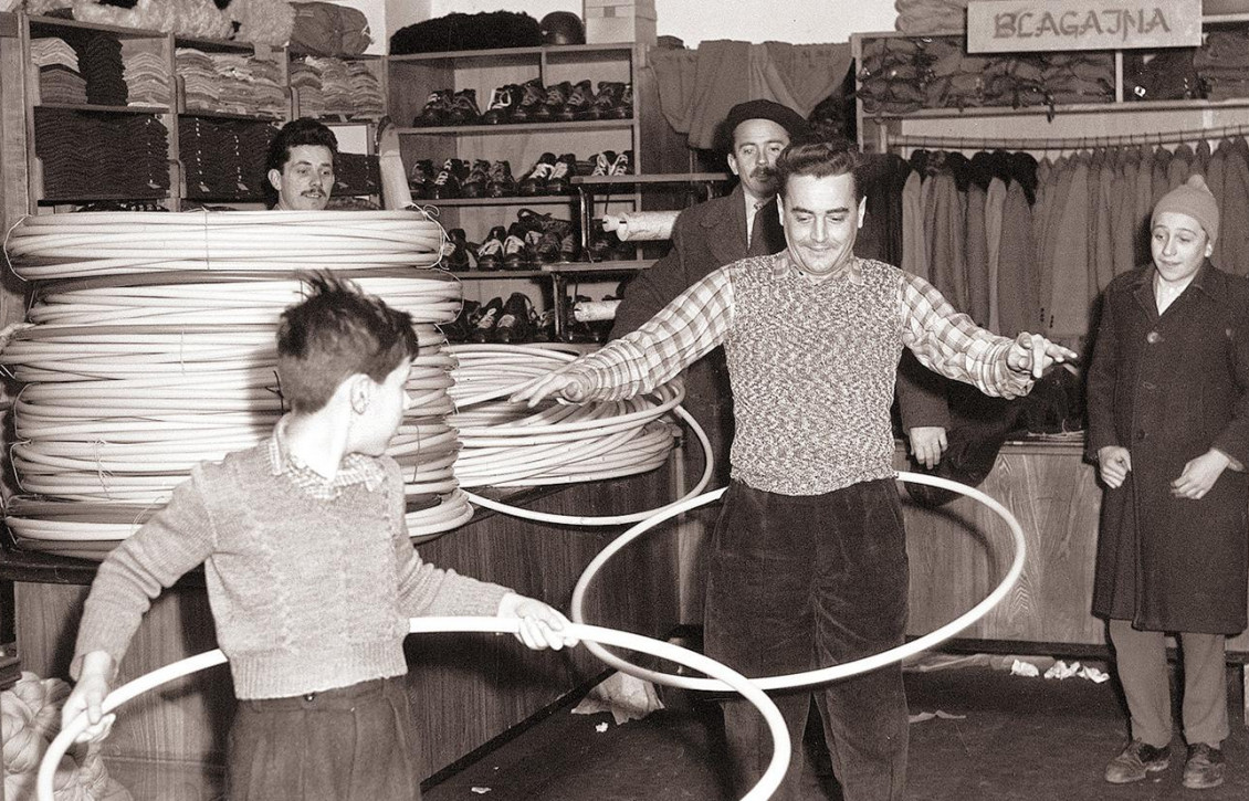 A store in Slovenia had just received its order of hula hoops in 1959. Who could resist trying one out? Photo, black and white, a man and a boy in a clothing store, swinging big hoops around their hips.