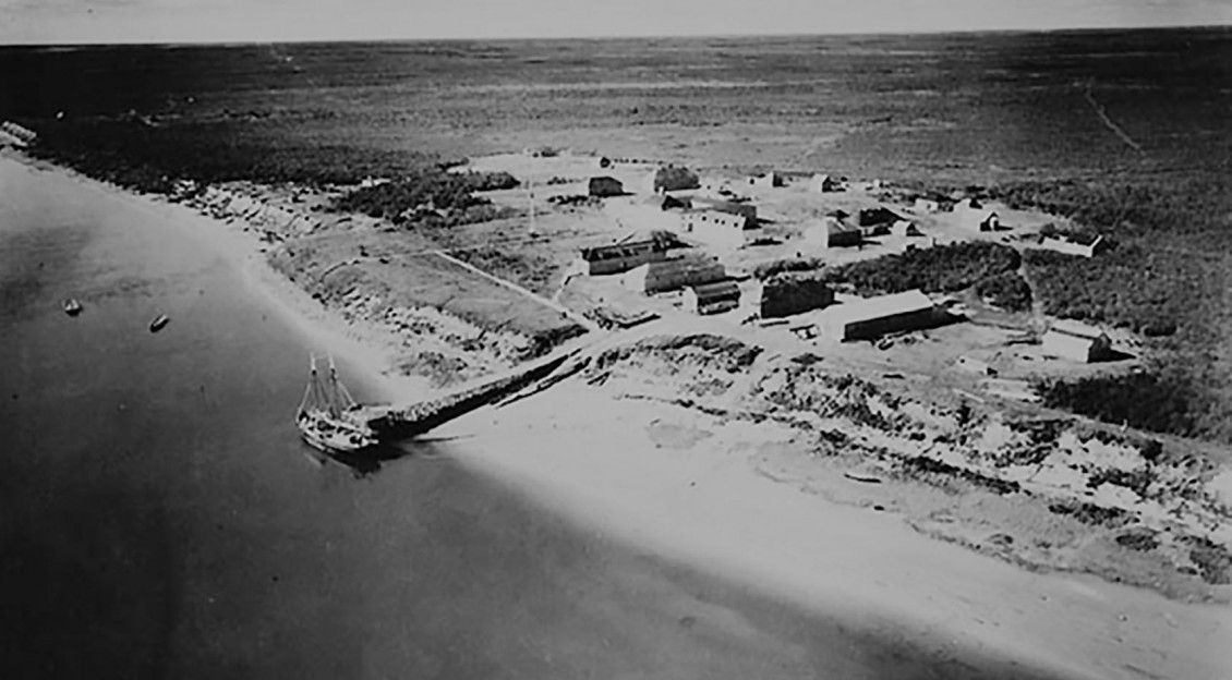 Photo, black and white, aerial view of vast empty land with military-style settlement on a bay.