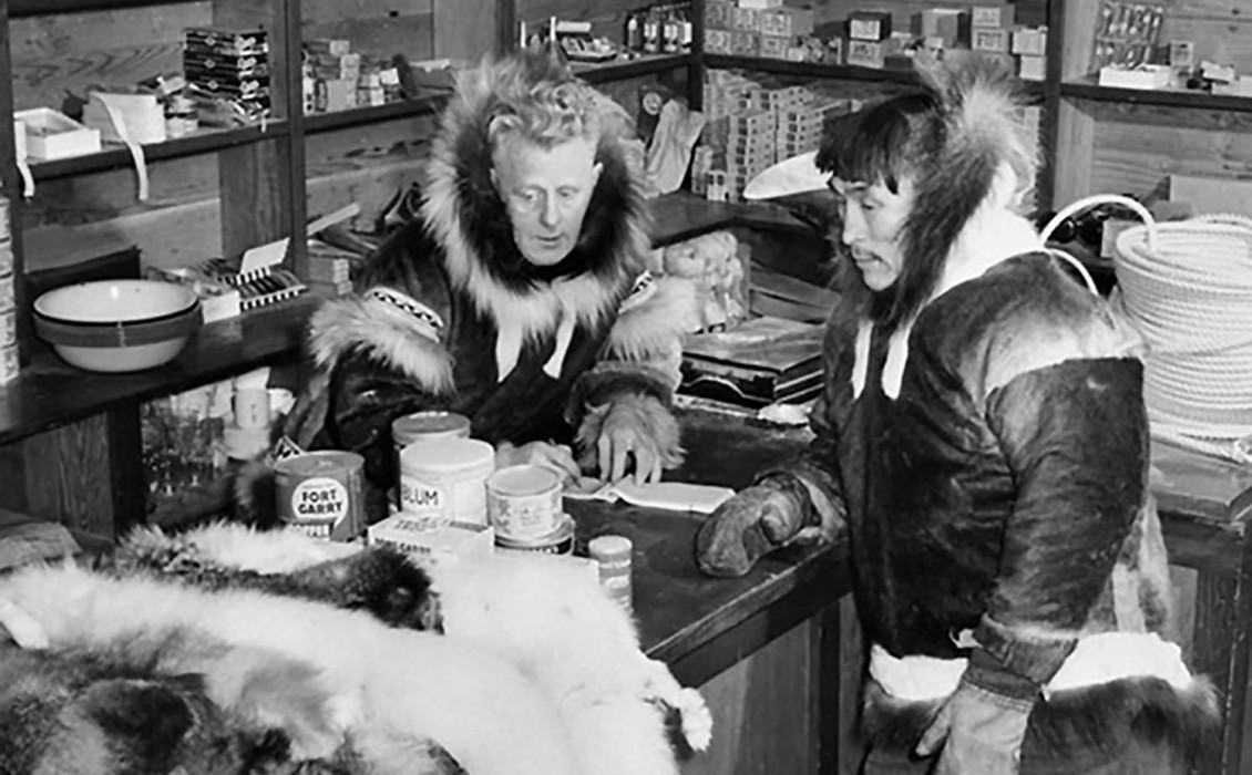 Photo, black and white, white man and Indigenous man wearing parkas in a room stocked with domestic supplies.