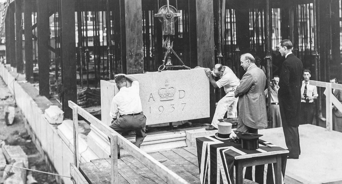 Photo, black and white, 2 men in formal wear watching 2 construction workers guide a block of stone hung from a crane.