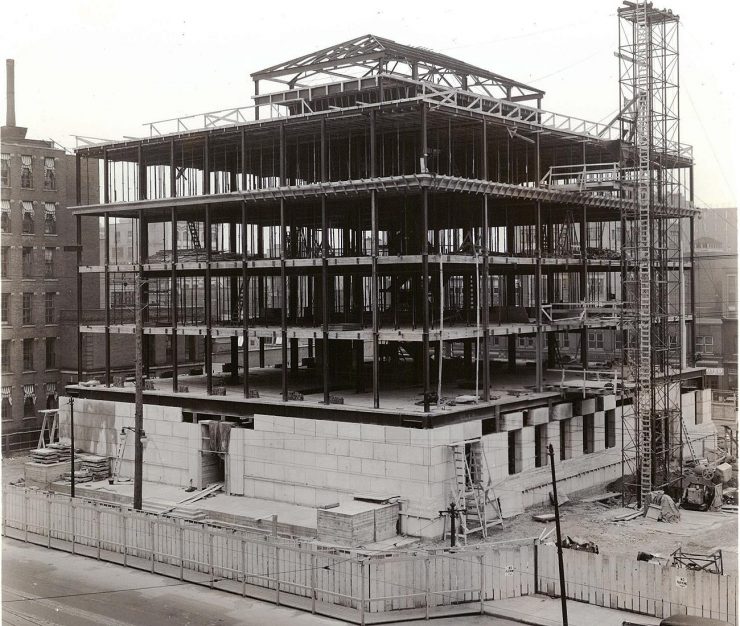 Photo, black and white, low, square building of iron girders with bottom-floor walls covered in stone.