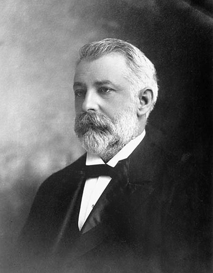 Photo, black and white, a bearded man with grey hair in a 19th century suit.