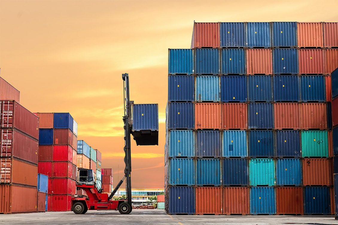 A photograph of colourful shipping containers stacked in neat rows. A lifting machine is holding a blue shipping crate. A warehouse building can be seen in the background.