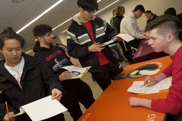 Teenagers, one seated at a table while others standing and holding clip boards consult with him.