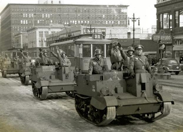 Photo, black and white, of a city street, with army vehicles full of men whose right arms are outstretched in salute.