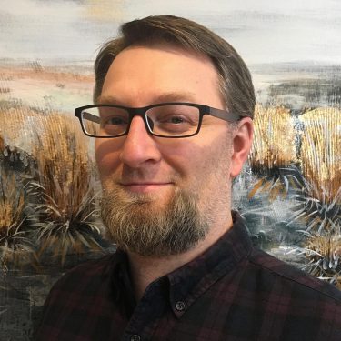 Photo, man, bearded, glasses, standing before a painting of a prairie landscape.