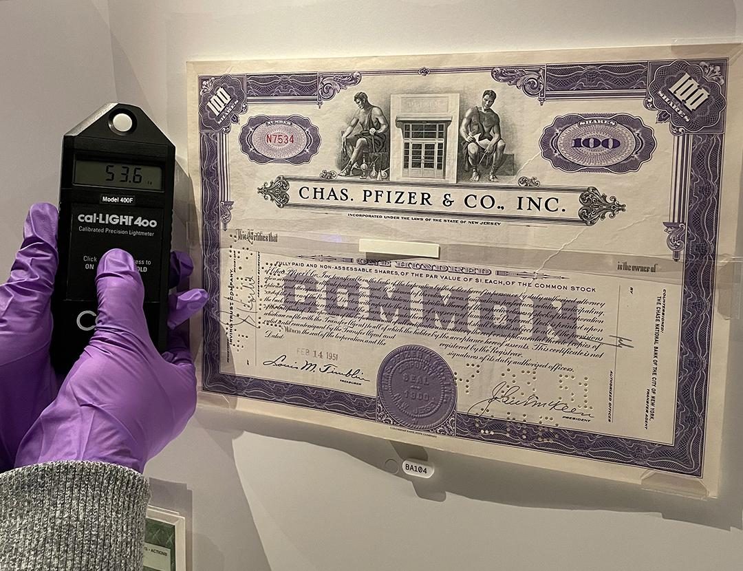A gloved hand holding a light measuring device in a display case of paper items.