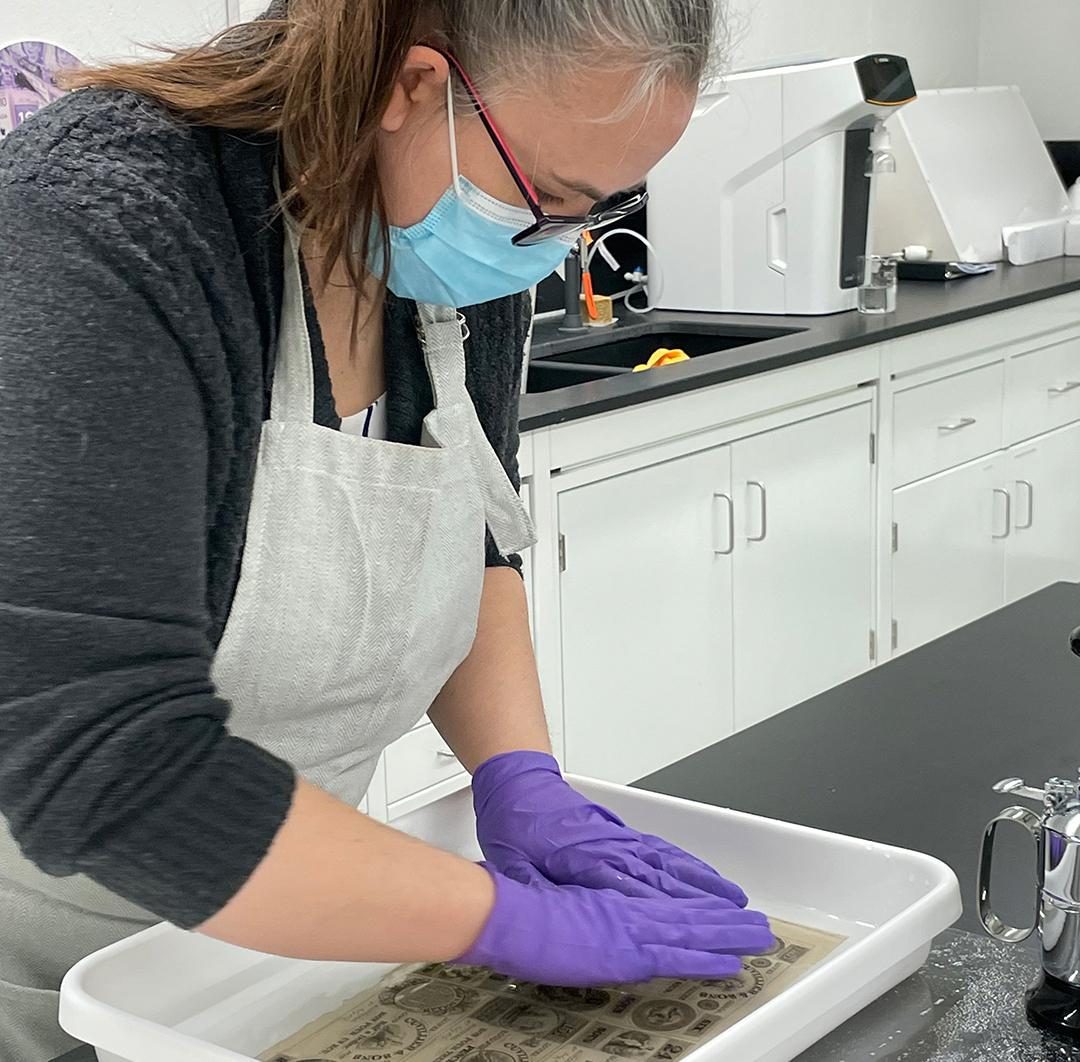 Woman in mask, gloves and apron soaking a paper document in a tray of water.