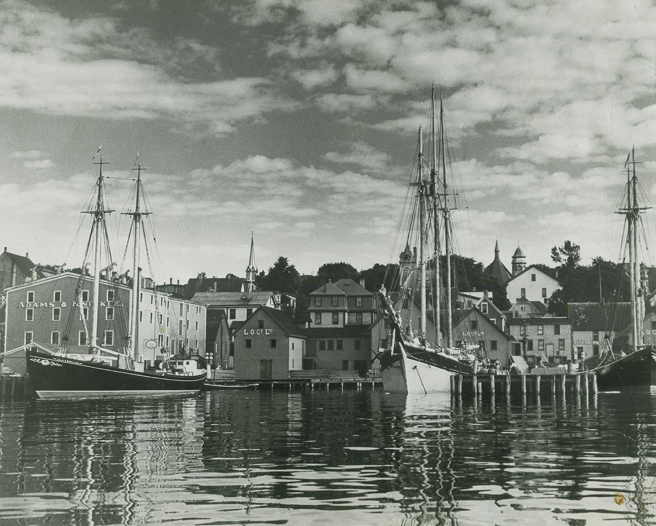 Photo, black and white, waterfront scene of wooden frame buildings and wharf with three sailing ships in dock.