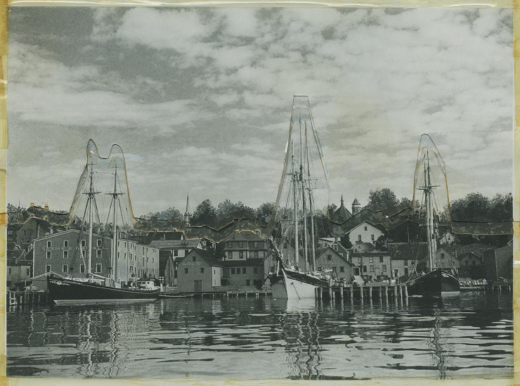 Photo, black and white, waterfront scene of wooden frame buildings and wharf with three sailing ships in dock.