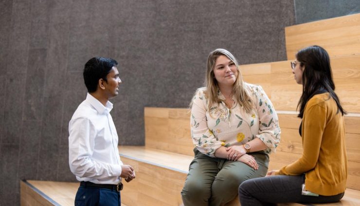Alt text: photo of three people in a conversation, two are sitting on wide bleachers.