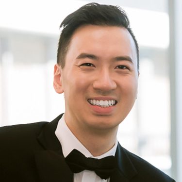 Photo, colour, smiling young man in a formal black tuxedo against a bright window.