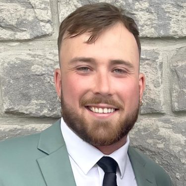 Photo, colour, smiling young man wearing a suit, standing before a stone wall.