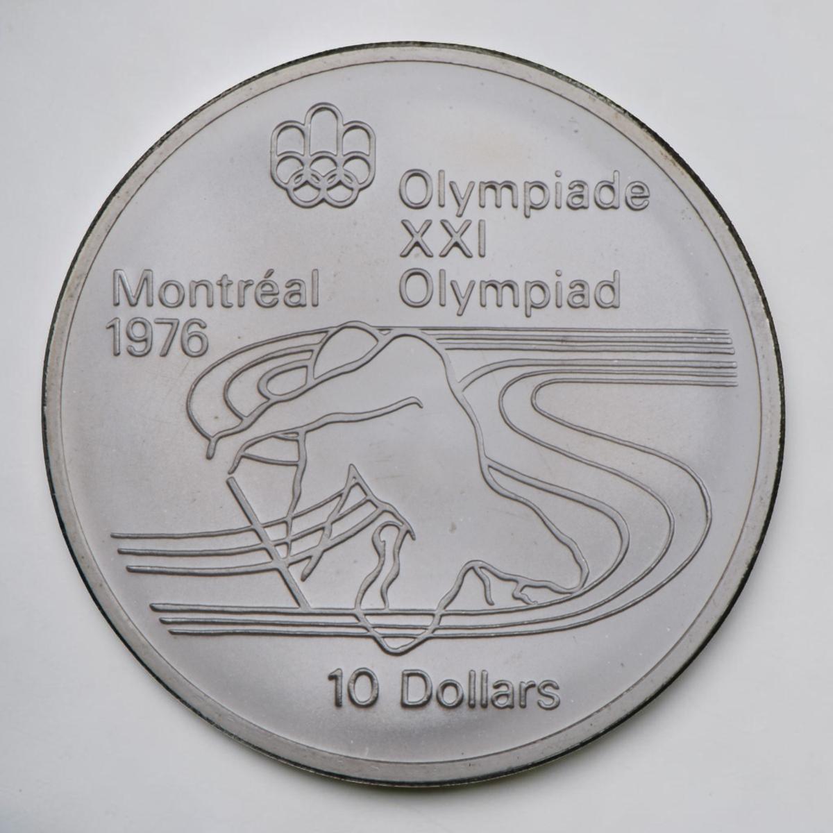 Coin, silver, stylized outline of an athlete kneeling and paddling in a patter of flowing lines.